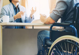 Une personne en fauteuil roulant discute avec un professionnel assis à un bureau, illustrant l’inclusion et l’apprentissage des personnes en situation de handicap en milieu professionnel.
