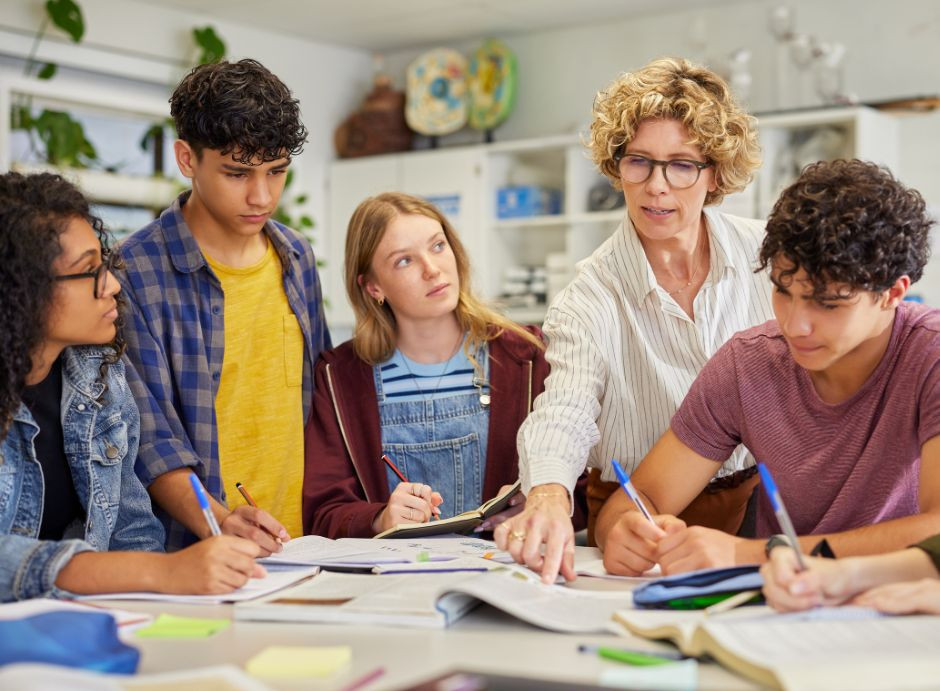 Cinq jeunes et une adulte, probablement une enseignante ou une formatrice, sont assis autour d’une table, en train de travailler ensemble sur des documents. L’image met en avant l’accompagnement éducatif, l’inclusion et le travail collaboratif pour aider les jeunes en difficulté à réussir.