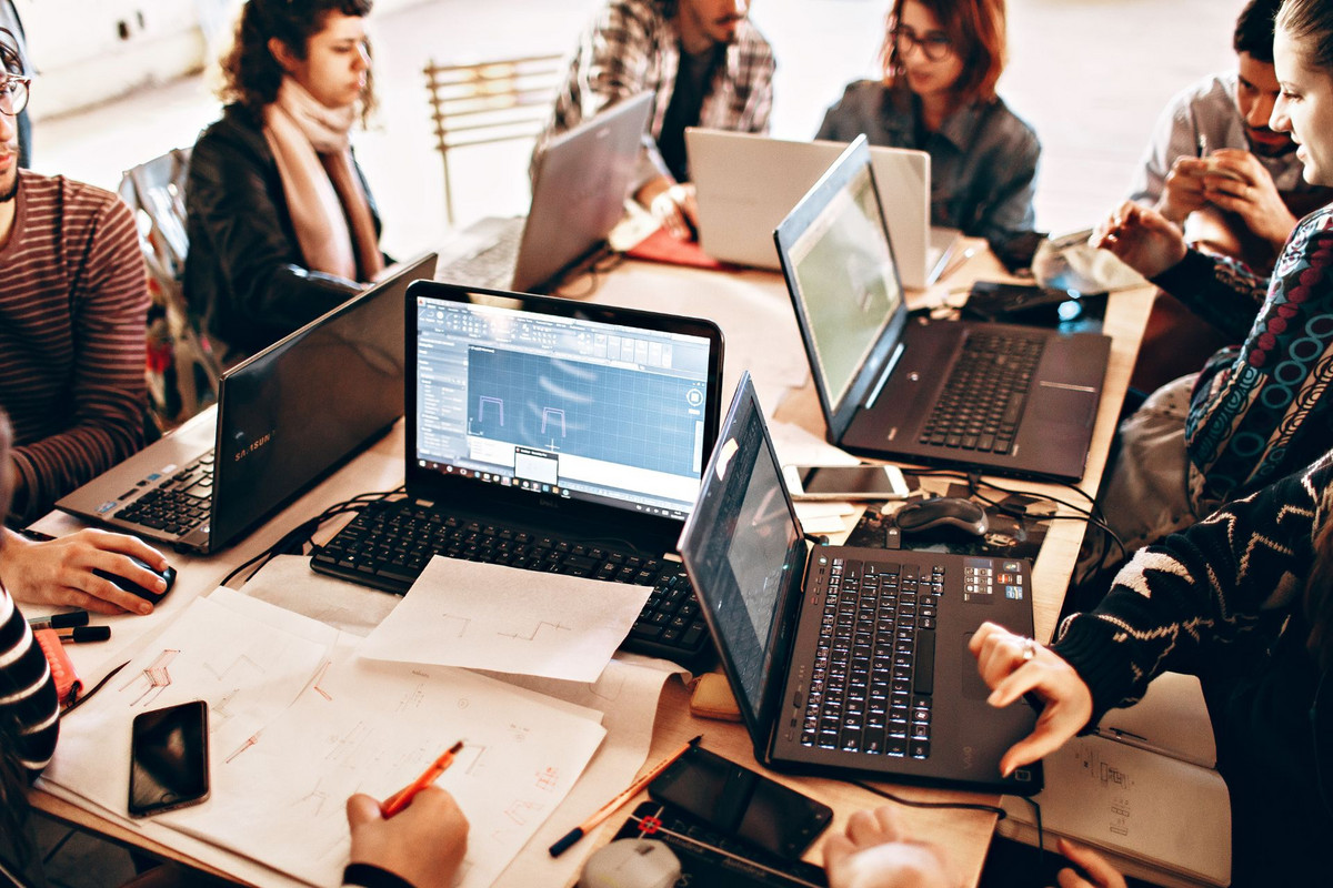L'apprentissage en action : collaboration et innovation Un groupe de jeunes et de formateurs travaille ensemble autour d'une table, entourés d'ordinateurs portables, de croquis et de documents. L'image illustre une séance de travail collaboratif et dynamique en apprentissage.