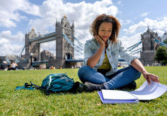 Jeune femme étudiante assise sur une pelouse devant Tower Bridge à Londres, avec un cahier et un sac à dos, illustrant l’apprentissage international et la mobilité des apprentis.