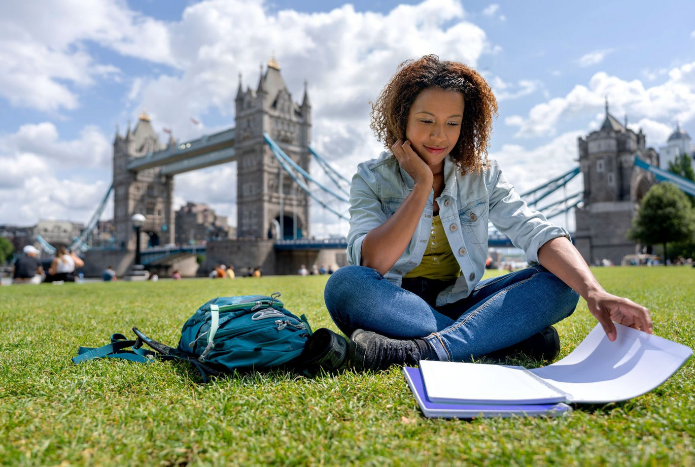 Jeune femme illustrant l’apprentissage international et la mobilité des apprentis Jeune femme étudiante assise sur une pelouse devant Tower Bridge à Londres, avec un cahier et un sac à dos, illustrant l’apprentissage international et la mobilité des apprentis.
