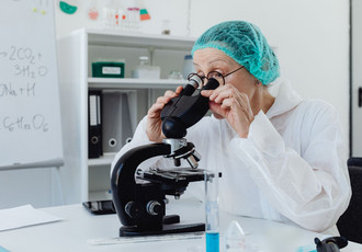 Femme scientifique en blouse blanche et charlotte bleue observant attentivement une lame au microscope dans un laboratoire.