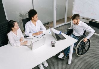 Trois personnes en réunion professionnelle autour d’une table : deux femmes et un homme en fauteuil roulant. Elles travaillent sur des ordinateurs portables et des documents, illustrant l’inclusion des personnes en situation de handicap dans le monde du travail grâce à des dispositifs comme l’alternance.