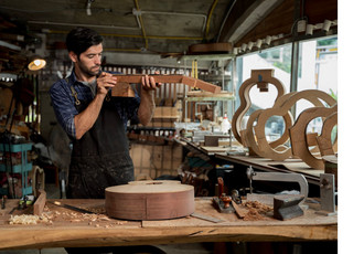 Artisan luthier travaillant le bois dans un atelier rempli d’outils et de pièces de guitare en construction, illustrant les métiers d’art et l’apprentissage traditionnel.