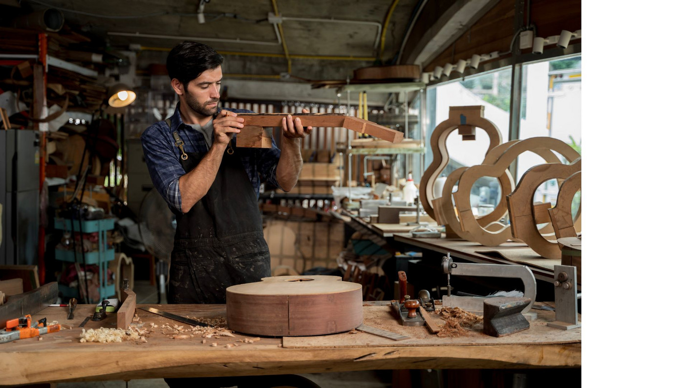 Artisan luthier travaillant le bois dans un atelier rempli d’outils et de pièces de guitare en construction, illustrant les métiers d’art et l’apprentissage traditionnel.