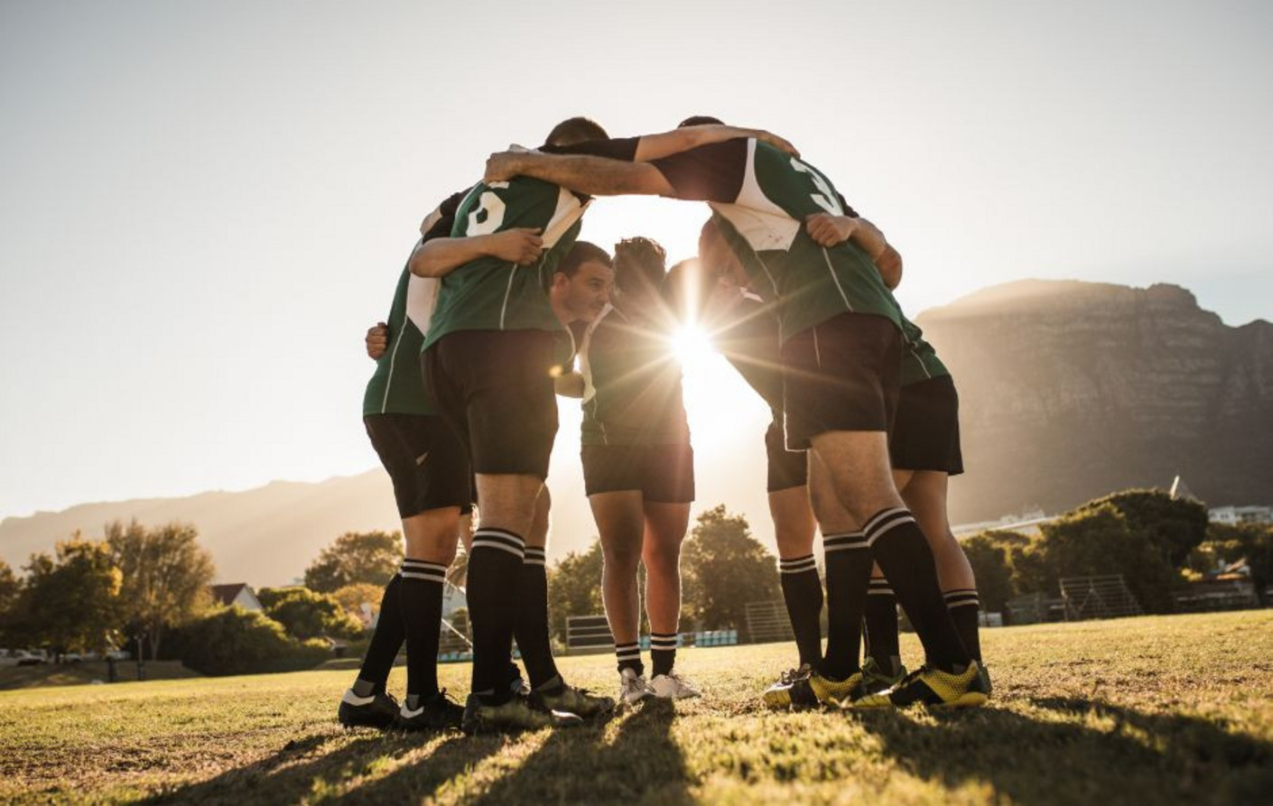 Cinq sportifs, vêtus de tenues d’entraînement, se serrent les épaules en cercle, sous un soleil couchant. Cette image représente la solidarité, la motivation et l’engagement nécessaires pour allier sport de haut niveau et formation en alternance.