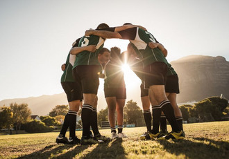 Cinq sportifs, vêtus de tenues d’entraînement, se serrent les épaules en cercle, sous un soleil couchant. Cette image représente la solidarité, la motivation et l’engagement nécessaires pour allier sport de haut niveau et formation en alternance.