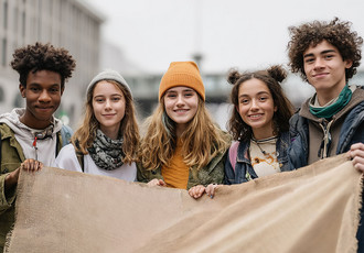 Cinq jeunes souriants, debout côte à côte en extérieur, tiennent ensemble une grande bannière en toile. Ils symbolisent l'engagement, la solidarité et la détermination des jeunes en apprentissage.
