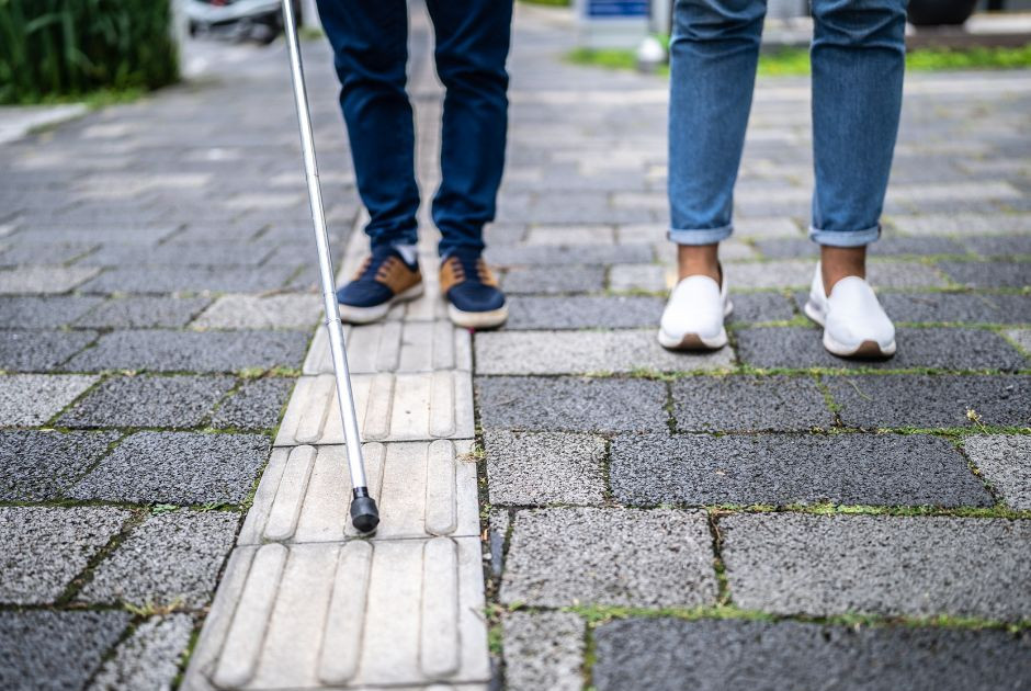 Accessibilité et autonomie : marcher ensemble vers une société inclusive Deux personnes marchent sur un trottoir pavé. La personne à gauche utilise une canne blanche pour se guider, tandis que la personne à droite l’accompagne. L’image met en avant l’inclusion et l’accessibilité pour les personnes en situation de handicap visuel.